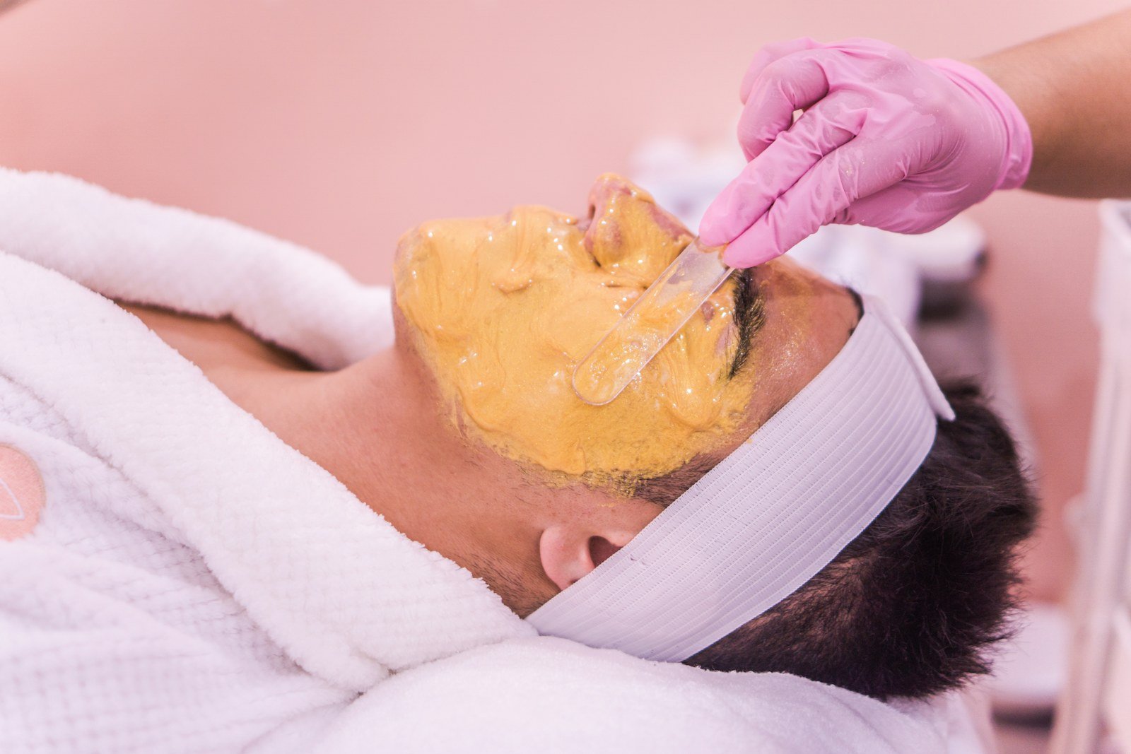 Esteticista de luvas aplicando máscara facial com espátula em cliente deitado na maca