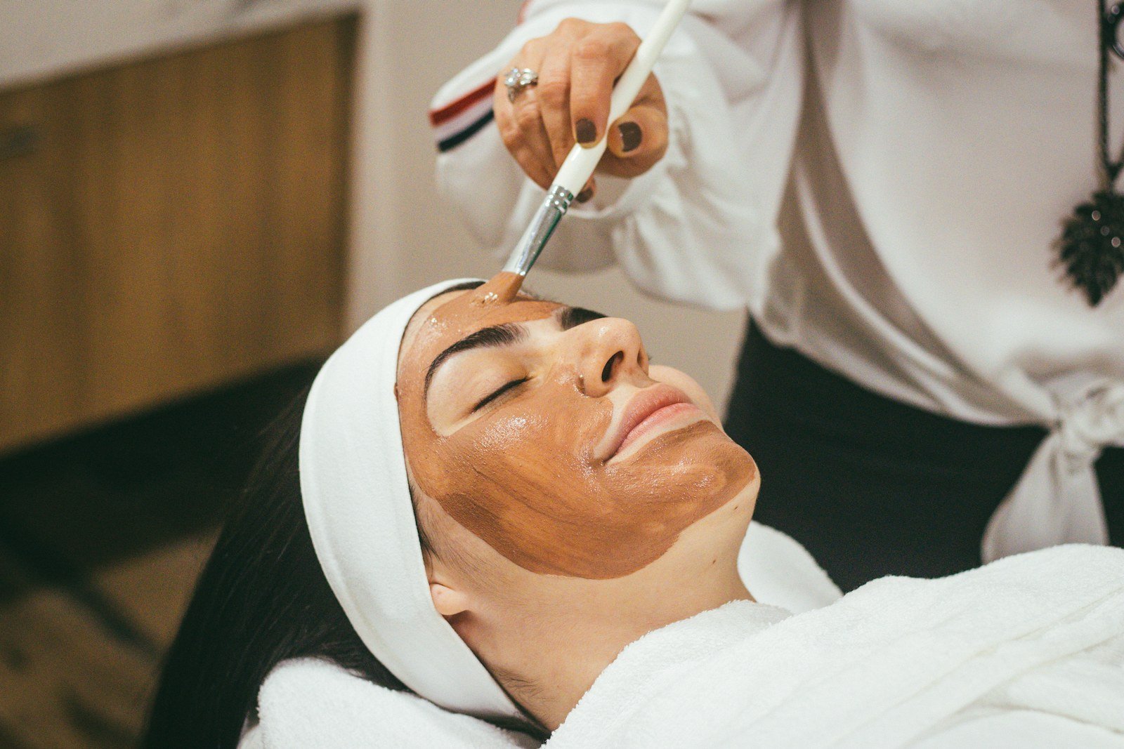 Spa de sobrancelha: profissional aplica argila com pincel na pele de uma cliente deitada, até a linha da sobrancelha